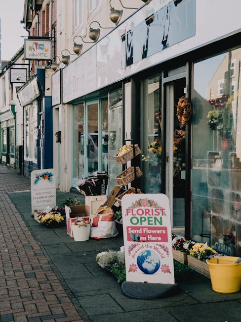 The image shows the storefront of a florist shop on a busy street, with large glass windows displaying floral arrangements inside. Outside the shop, various gardening supplies and potted plants are ar